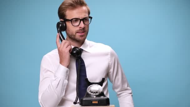 attractive businessman answering an old telephone, talking with somebody and getting mad, arguing and screaming then slamming the phone on blue background - Footage, Video