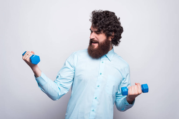 Фото бородатого чоловіка в повсякденному одязі синьої сорочки, що працює з маленькими гантелями
. - Фото, зображення