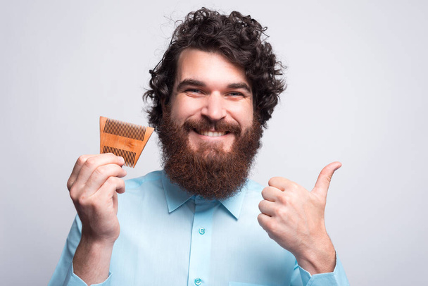 Φωτογραφία του γενειοφόρου hipster άνθρωπος κρατώντας βούρτσα μαλλιών και δείχνει τον αντίχειρα μέχρι. - Φωτογραφία, εικόνα