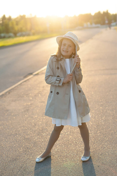 A little girl in a beige raincoat stands in the setting sun. Family walk in the Park. - Zdjęcie, obraz