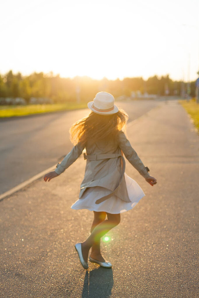 Маленька дівчинка в бежевому плащі стоїть на заході сонця. Сімейна прогулянка в парку
. - Фото, зображення