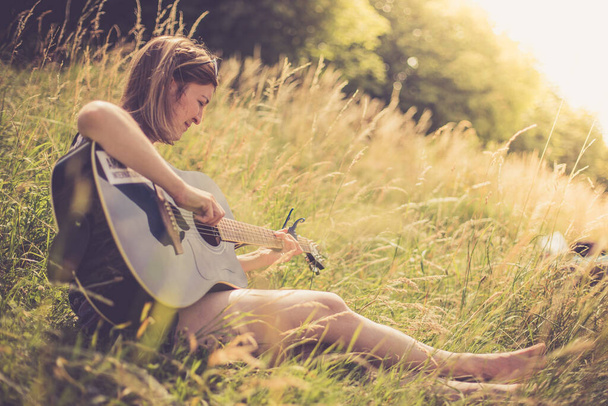 Beautiful young girl is sitting at a river and playing her western guitar - Foto, afbeelding