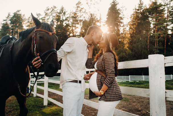 a pregnant girl in a hat and a man in white clothes stand next to horses near a white fence.Stylish pregnant woman with a man with horses.Married couple - Foto, Imagem