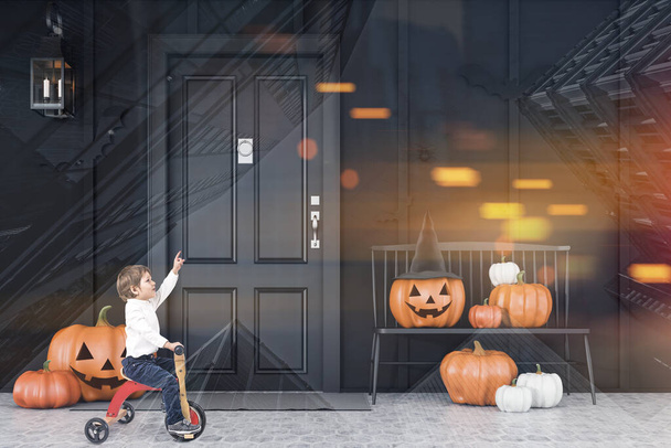 Little boy on tricycle riding near black bench with carved pumpkins in front of modern house with black doors and walls and halloween decorations. Toned image double exposure - 写真・画像