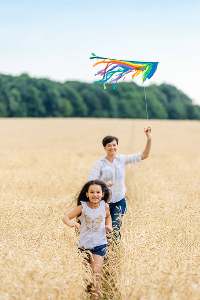 Mother and daughter run in a wheat field with a kite in the summer. Happy childhood. - Photo, image