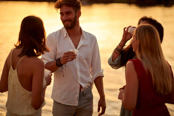Young handsome man flirting and drinking with the girls on a party on the lake with a beautiful sunset - Photo, image