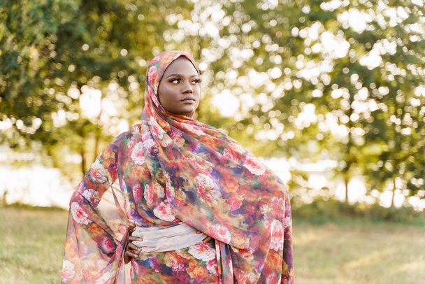 Μουσουλμάνα γυναίκα Αφρικανική εθνότητα φορούσε παραδοσιακή πολύχρωμη μαντίλα κοιτάζει δεξιά πλευρά στο πράσινο λιβάδι. Αραβικά ισλαμικά θρησκευτικά ρούχα. Ένα όμορφο μαύρο κορίτσι χαμογελάει - Φωτογραφία, εικόνα