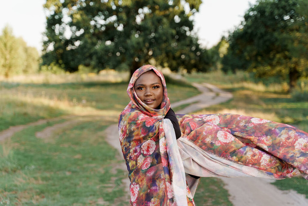 Κομψό ελκυστικό μουσουλμανικό κορίτσι σε μοντέρνα παραδοσιακά ρούχα, γυναίκα με παραδοσιακή μαντίλα. Πολιτισμοί όλου του κόσμου. Αφρικανικός τρόπος ζωής στο πράσινο λιβάδι. - Φωτογραφία, εικόνα