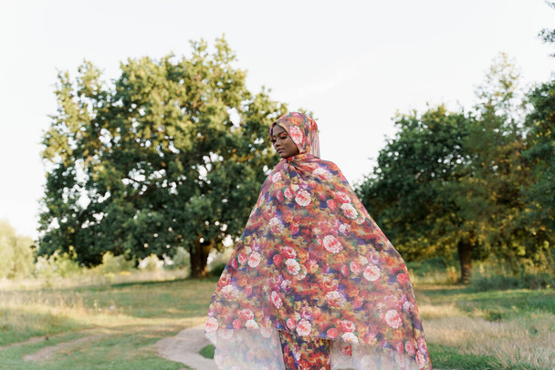 Stylowa atrakcyjna muzułmańska dziewczyna w modnej tradycyjnej odzieży, kobieta w tradycyjnym hidżabie. Dziewczyna wygląda na lewą. Kulturą całego świata. Afrykański styl życia na zielonej łące. - Zdjęcie, obraz