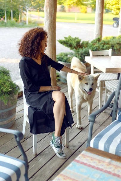 Vue verticale d'une jeune belle femme aux cheveux bouclés, vêtue d'une robe noire, perlée sur la terrasse jouant avec son chien. - Photo, image