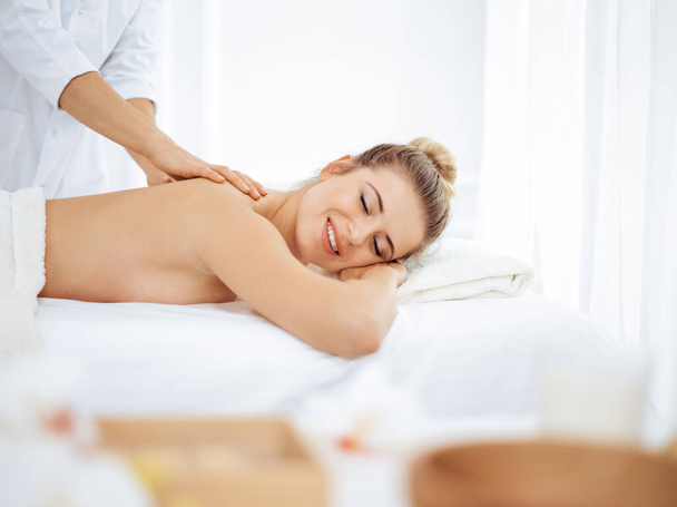 Young and blonde woman enjoying massage of back in spa salon. Beauty concept - Photo, Image