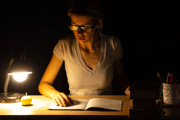 une fille s'assoit à une table à la lumière d'une lampe dans une pièce sombre et fait des devoirs, des devoirs, des devoirs - Photo, image