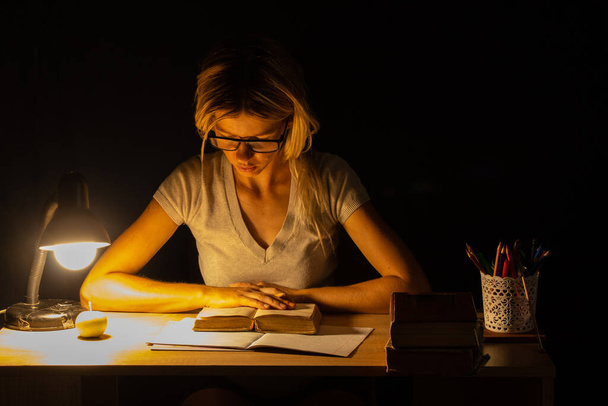 una ragazza siede a un tavolo alla luce di una lampada in una stanza buia e fa i compiti, i compiti, i compiti - Foto, immagini