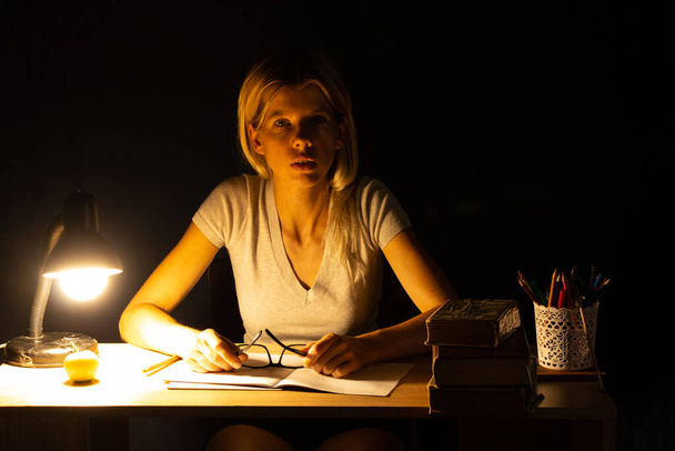 ένα κορίτσι κάθεται σε ένα τραπέζι δίπλα στο φως μιας λάμπας σε ένα σκοτεινό δωμάτιο και κάνει την εργασία, την εργασία, την εργασία - Φωτογραφία, εικόνα