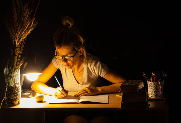 dziewczyna siedzi przy stole przy lampie w ciemnym pokoju i odrabia pracę domową, pracę domową, pracę domową - Zdjęcie, obraz