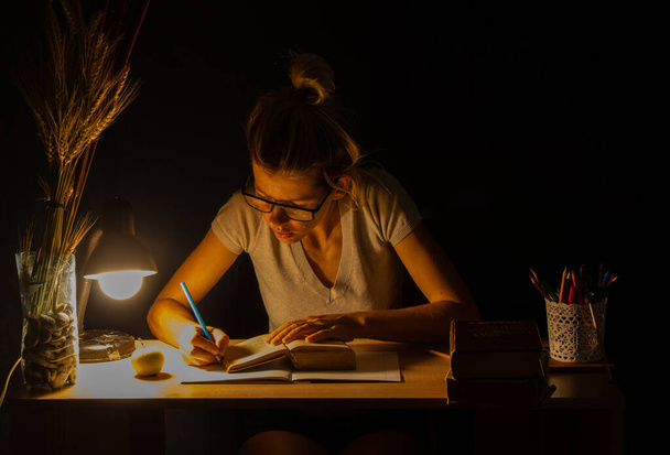 tyttö istuu pöydässä lampun valossa pimeässä huoneessa ja tekee kotitehtäviä, kotitehtäviä, kotitehtäviä - Valokuva, kuva