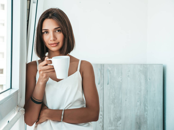 Portrait of beautiful girl dressed in white pajamas. Carefree woman holds cup and drinking coffee. Sexy model enjoying her morning breakfast at balcony. Female in her thoughts  - Photo, Image