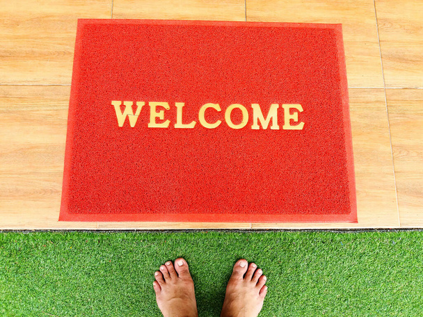 Top view of feet on green glass field and red foot towel, rug or carpet with welcome wording and copy space. Flat lay of part body and colorful background. - Photo, Image