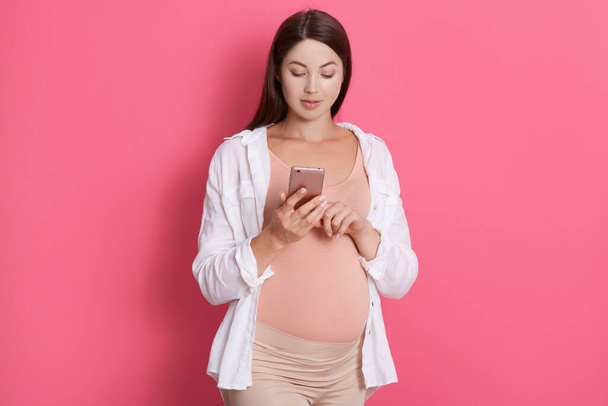 Beautiful pregnant woman dialing number, holding mobile phone in hands, looking at device's screen, standing with concentrated look isolated over pink background. - Fotografie, Obrázek