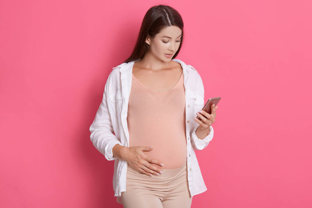 Young pregnant woman with dark hair wearing white shirt holds smart phone in her hand and looks at camera isolated over pink background, texting with doctor and touches belly. - Valokuva, kuva