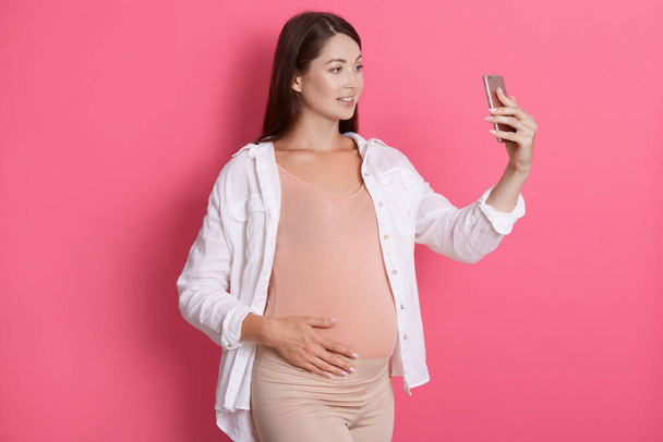 Young pregnant woman takes selfie photo with smart phone or having video call against pink wall, happy expectant mother using wireless internet for online communication. - Foto, afbeelding