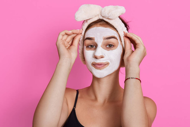 Young lady wearing sleeveless t shirt and headband, looking at camera with cosmetic mask on face, doing beauty procedures in morning at home. - Foto, imagen