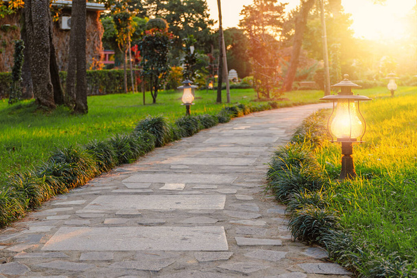 Sentiero lastricato in pietra nel parco tra prato verde e alberi. Lampade accese al tramonto. Batumi, Georgia. - Foto, immagini