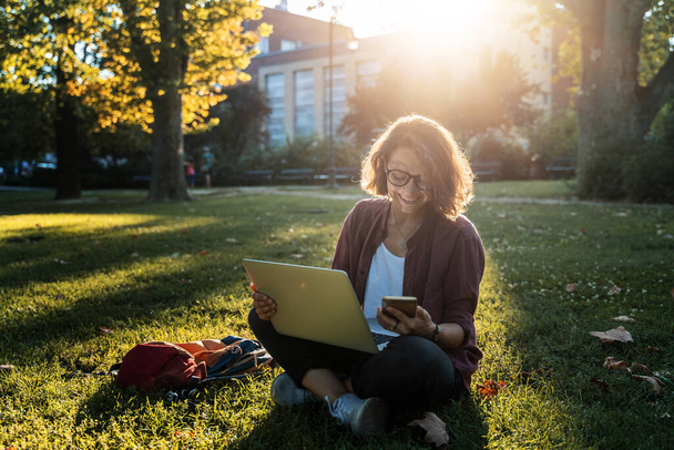 Young businesswoman student working on her notebook in park. Freelancer female sitting on grassy lawn using laptop with mobile phone on grass. - Fotografie, Obrázek