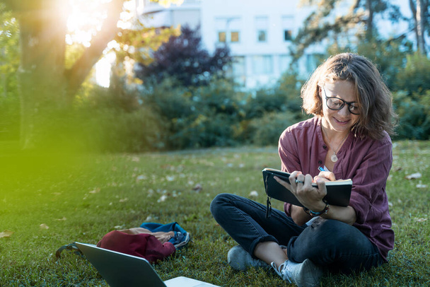 Young curly woman student sits on the grass and writes with a pen in a notebook sitting in front of a laptop in park - Photo, Image