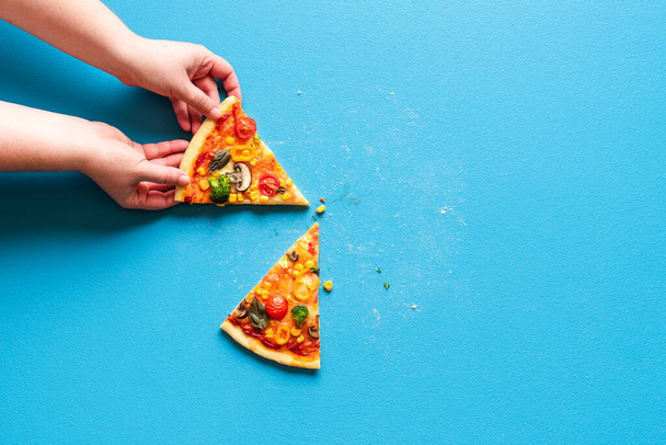 A mão da mulher agarra uma fatia de pizza vegetariana. Vista acima com fatias de pizza primavera em uma mesa de cor azul. - Foto, Imagem