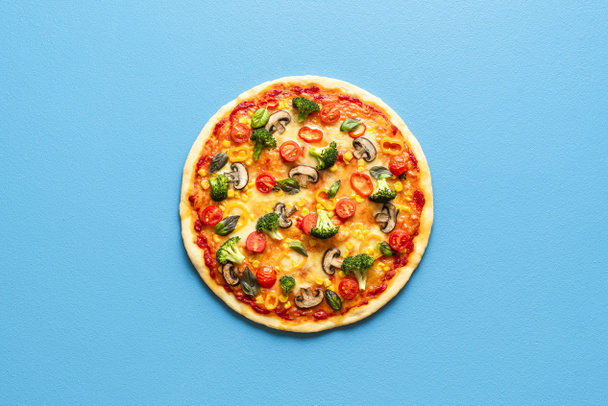 Bovenaanzicht met een hele pizza primavera geïsoleerd op een blauw gekleurde achtergrond. Vegetarische pizza met broccoli, champignons, tomaten, maïs en peper. - Foto, afbeelding