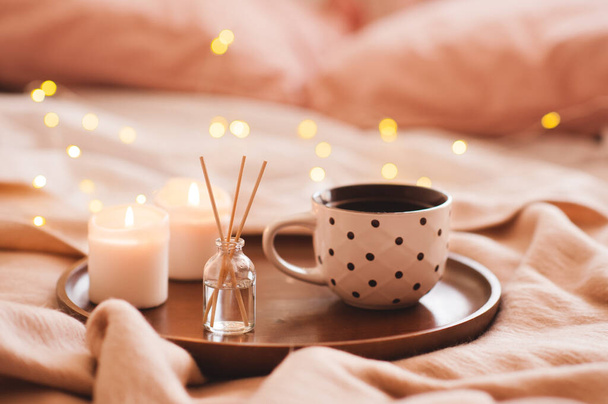 Cup of coffee with home aroma stickes and burning candles in bed closeup over glowing christmas lights. Winter holiday season.  - Photo, Image