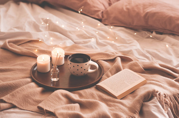 Cup of coffee with aroma sticks, open book and burning candles on wooden tray in bed on knitted blanket closeup. Winter season. Cozy atmosphere concept. - Photo, Image