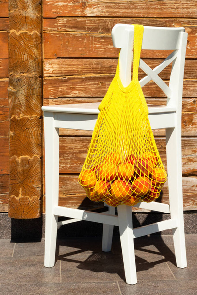 Сетчатый мешок или веревочный мешок с спелыми персиками на спинке белого стула. Ноль отходов. - Фото, изображение