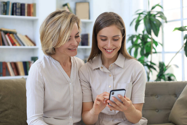 Middle aged mother and adult daughter hugging, using phone together, watching video or photos, sitting on cozy couch at home - Fotografie, Obrázek