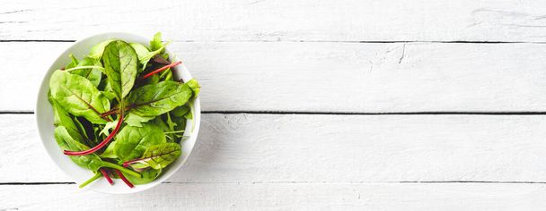 Overhead shot of fresh green salad with spinach, arugula and beetroot leaves in bowl on white wooden background with copyspace. Banner - Photo, image