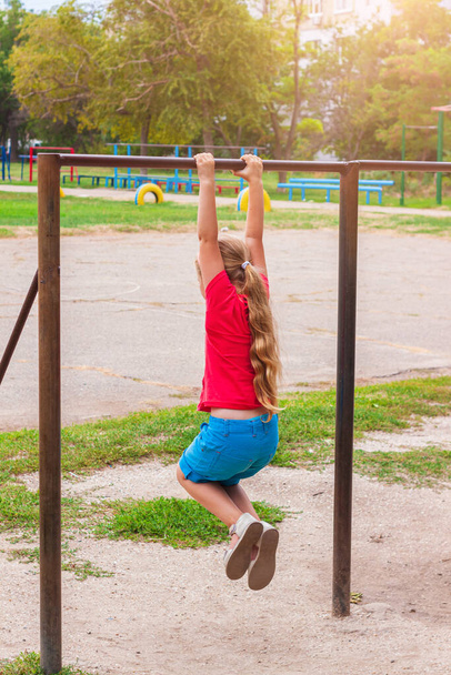 Κορίτσι κρέμεται σε οριζόντια γραμμή. Παιδί κάνει ασκήσεις κίνησης και παίζει σε παιδική χαρά εξωτερική. Υγιής αθλητική δραστηριότητα αναψυχής στον καθαρό αέρα. Ευτυχισμένη παιδική ηλικία. Παιδική σωματική άσκηση σε αθλητικό στάδιο. - Φωτογραφία, εικόνα