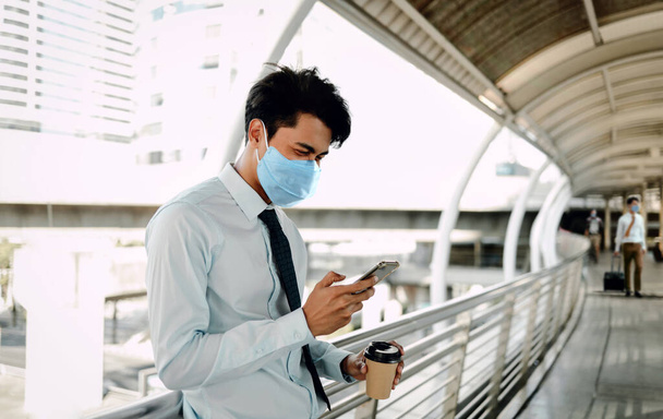 Nuori aasialainen liikemies yllään kirurginen naamio ja käyttämällä älypuhelinta kaupungissa. Terveydenhuolto uudessa normaalissa elämäntavassa käsite - Valokuva, kuva