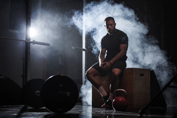 Muscular athlete with weightlifting equipment. Crossfit trainer in a fitness studio. Smoke background. - 写真・画像