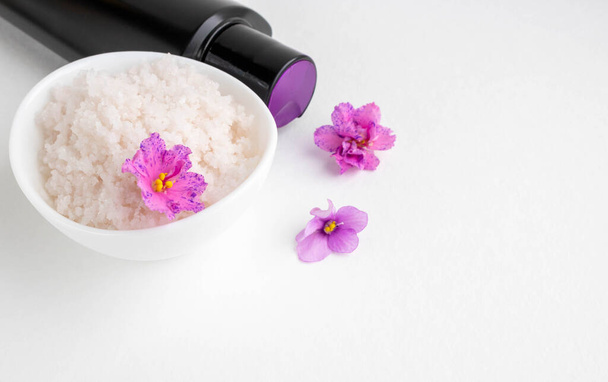 Овальная белая миска с розовой солью стоит на белом фоне рядом с ней черная бутылка и фиолетовые цветы. - Фото, изображение