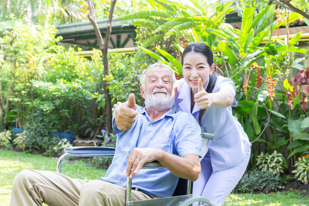 Egy nővér gondját viseli egy idősebb férfinak, és felhúzza a hüvelykujját a kerekesszékben a kertjében, otthon. - Fotó, kép