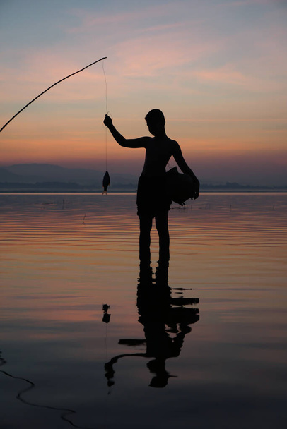 Gölün kenarında, Asyalı balıkçı gün doğumunda balık tutmak için olta kullanıyor. - Fotoğraf, Görsel