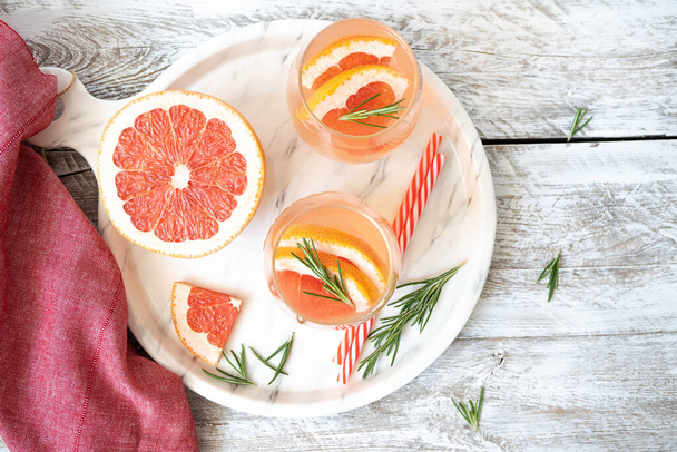 Two glasses of citrus lemonade with grapefruit wedges and rosemary stand on a white tray on a light wooden table. Nearby are grapefruit wedges, striped cocktail tubes and fresh rosemary. Top view - Foto, Imagem