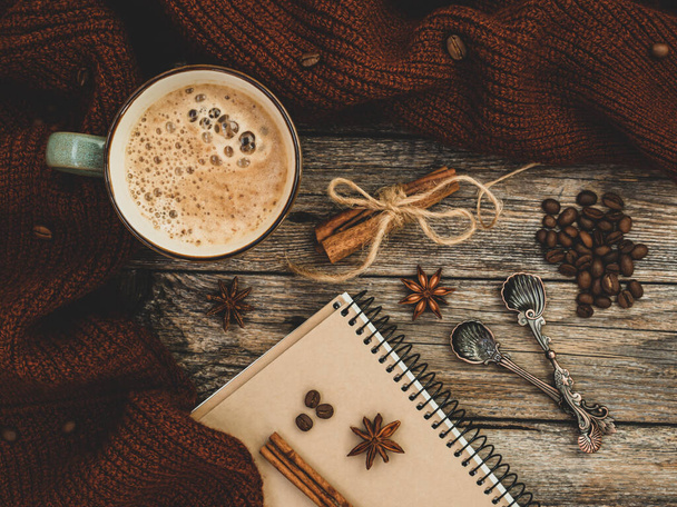 Útulný pletený hnědý svetr a aromatický šálek kávy, skořice, anýzové hvězdy, lžíce, kávová zrna a poznámkový blok na dřevěném pozadí. Podzimní nebo zimní kompozit. Nálada, životní styl, zátiší. - Fotografie, Obrázek