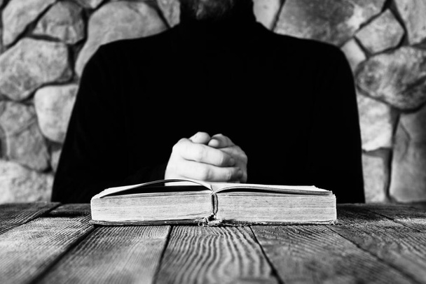 a man in black clothes in front of an open old book the Bible or the Koran, the concept of prayer, study, witchcraft. selective focus, black and white photo - Fotó, kép