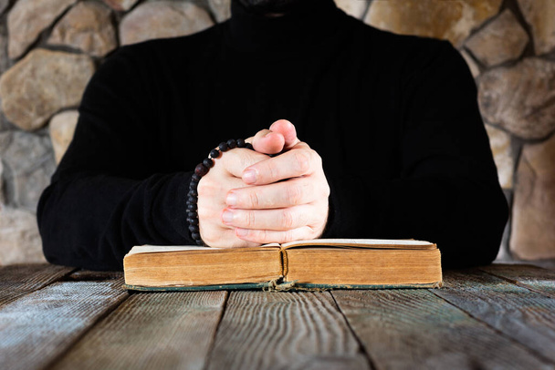 a man in black clothes with a rosary in his hands in front of an open old book the Bible or the Koran, the concept of prayer - Photo, Image