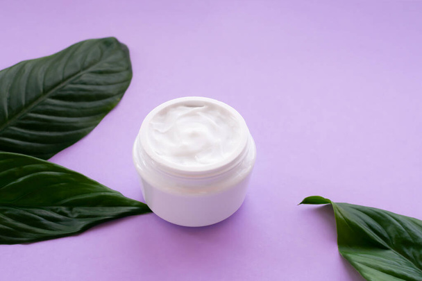 Soft white cream in jar with bright green leaves on purple background. Spa treatment and procedure at home. - Фото, изображение