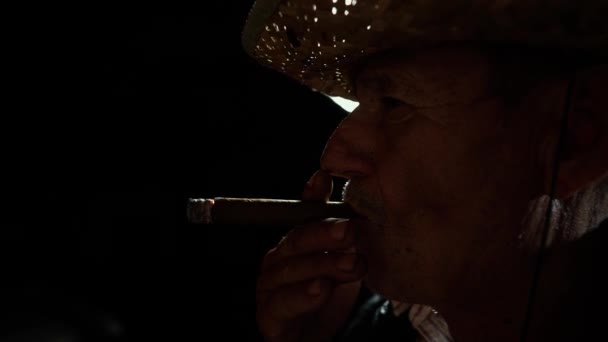 De man rookt een sigaar. Een oudere boer met een strohoed rookt een sigaar buiten de ranch. Dikke tabaksrook. Zelfverzekerde señor weerspiegelt zich in sigarettenrook. Een oude cowboy in een leren vest. - Video