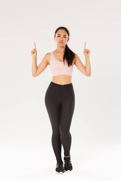 Sport, gym and healthy body concept. Full length of unamused and skeptical fitness girl, asian female athlete in sportswear complaining, sulking displeased and pointing fingers up, white background - Foto, imagen