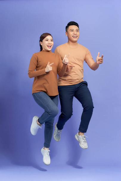 Excited young Asian couple man and woman jumping and pointing with happy and fun over purple background - Foto, imagen